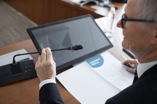 A senior businessman using a microphone during a conference meeting with documents and a monitor, illustrating an AI industry executive addressing safety concerns.